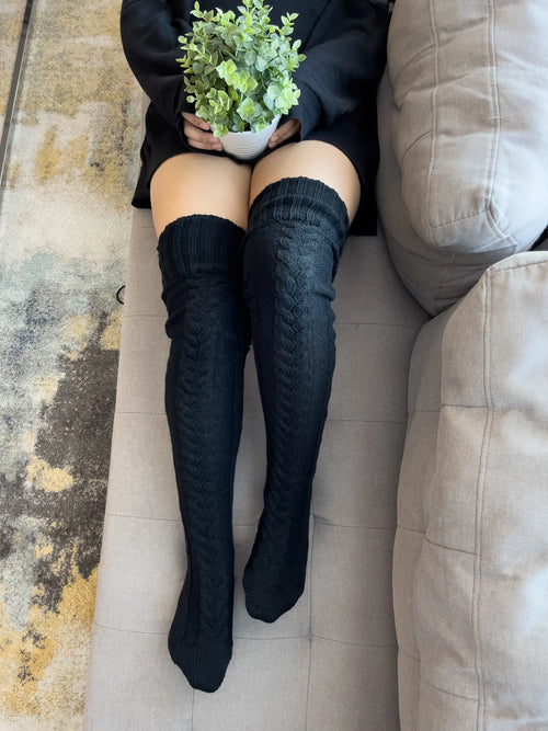 Black cable knit thigh high socks, worn on a sofa with an oversized sweater
