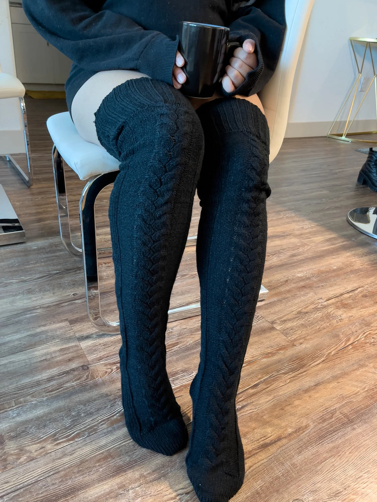 Black cable knit thigh high socks, worn sitting on a chair with a coffee mug
