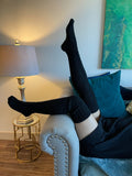 Black cable knit thigh high socks, leg raised in a cozy living room