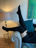 Black cable knit thigh high socks, leg raised in a cozy living room