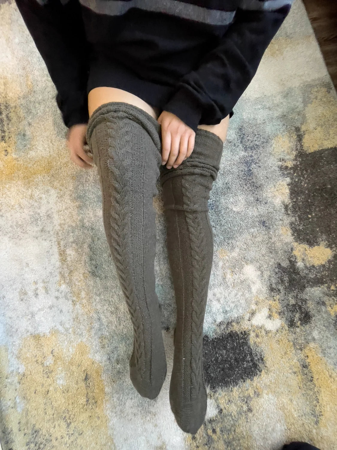 Charcoal cable knit thigh high socks, worn with a black sweater on a rug