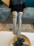 Gray cable knit thigh high socks, worn with a striped sweater on a sofa