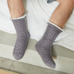 Gray men's fleece-lined slipper socks, worn sitting on a bed